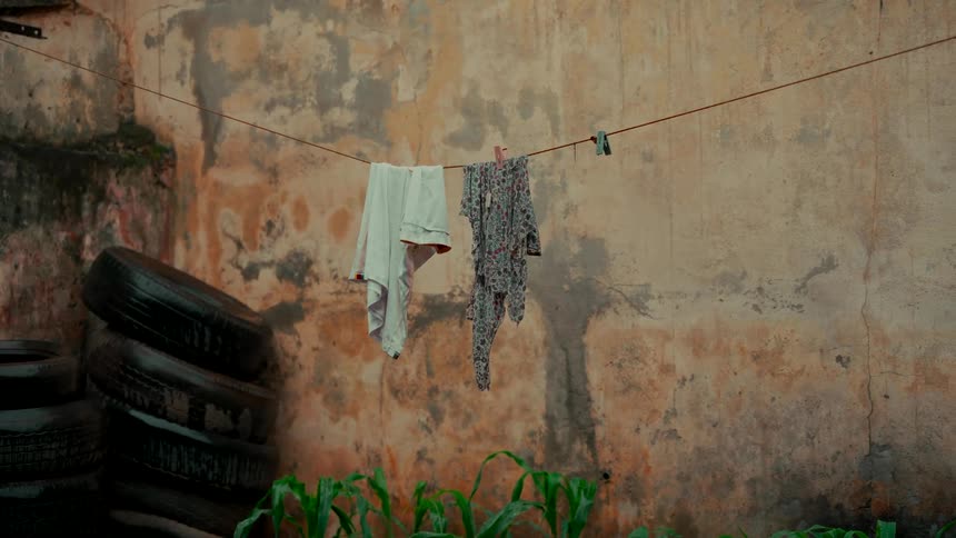 Laundry hangs on a clothesline to dry in a rustic backyard against a weathered wall next to a stack of old tires