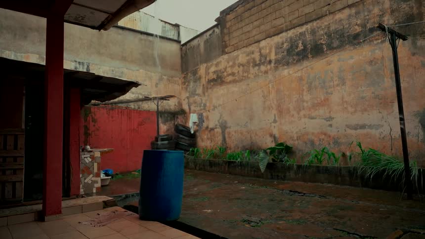 Rain falls steadily into a dilapidated urban courtyard, collecting in a large blue barrel under a broken gutter