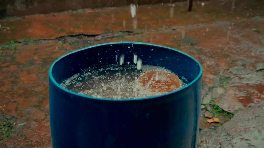 Close-up slow motion video of raindrops splashing as they collect in a blue water barrel on a wet patio