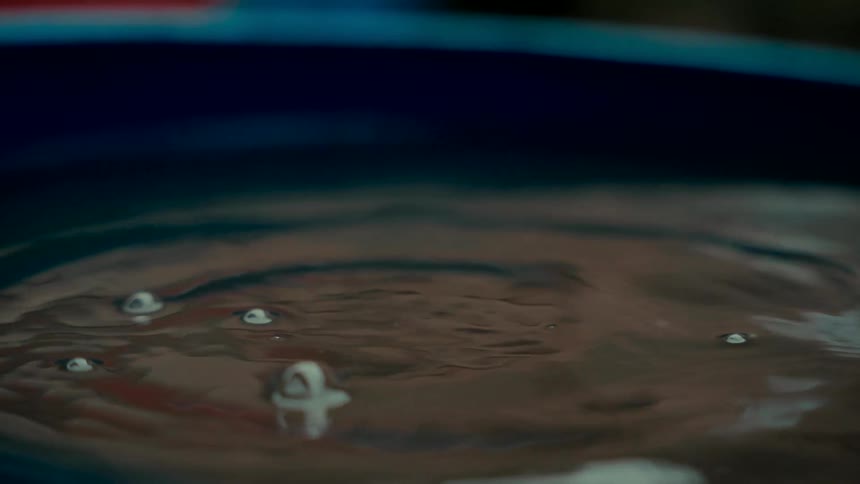 A close-up slow-motion shot captures a single water droplet hitting a calm surface, creating a beautiful crown splash and sending ripples across the dark water