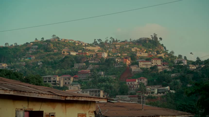 Wide shot of a densely populated village sprawling across a lush, green hillside in Freetown, Sierra Leone during a warm sunset
