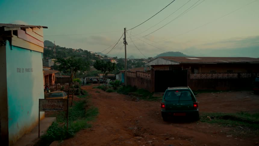 A quiet dirt road runs through a humble hillside village at dusk, with a small car parked beside a weathered building