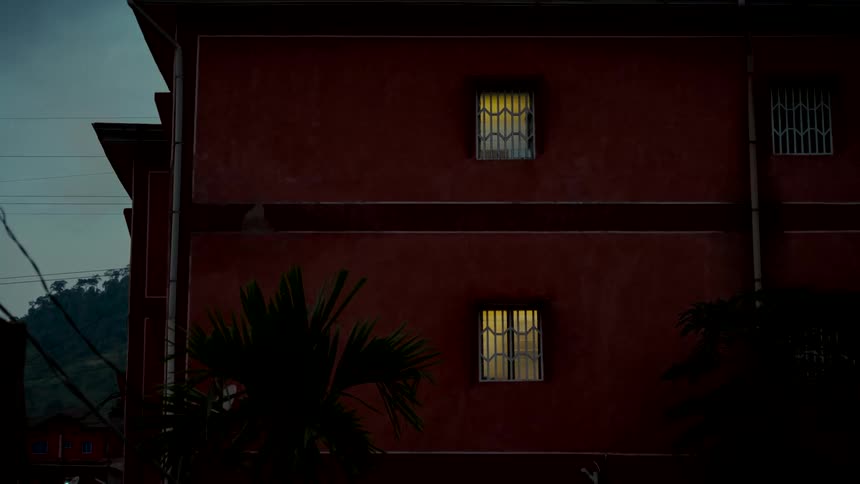 A still shot of a dark red apartment building at dusk, where two windows glow with warm yellow light