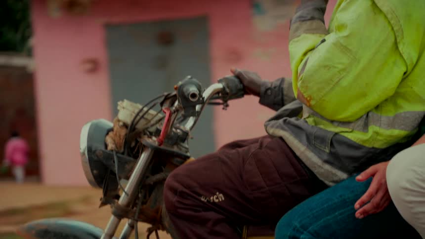 A close-up slow-motion shot of two people riding a weathered motorcycle through a dusty village street