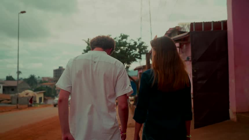 A slow-motion tracking shot from behind a young couple as they walk down a dusty street through a small town in Africa