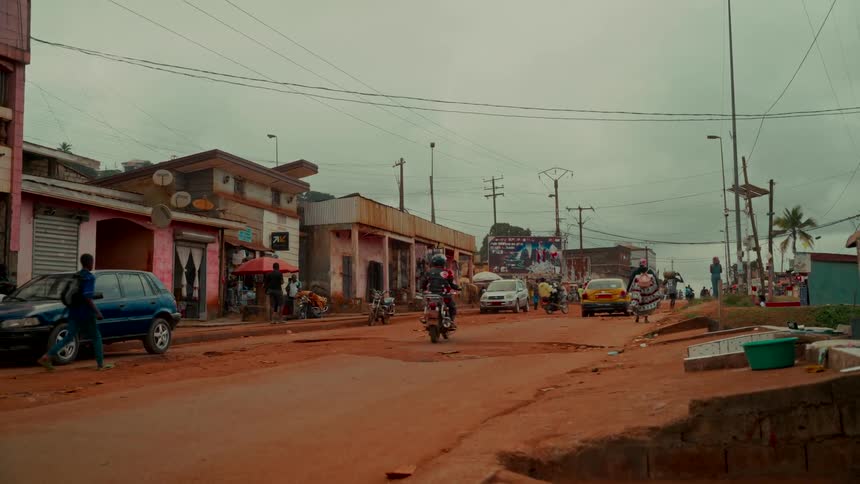 A motorcycle drives along a busy red dirt road through a town in Africa on a cloudy day