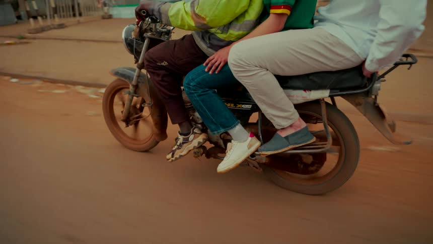 A tight tracking shot follows three men sharing a ride on a motorcycle down a dusty road