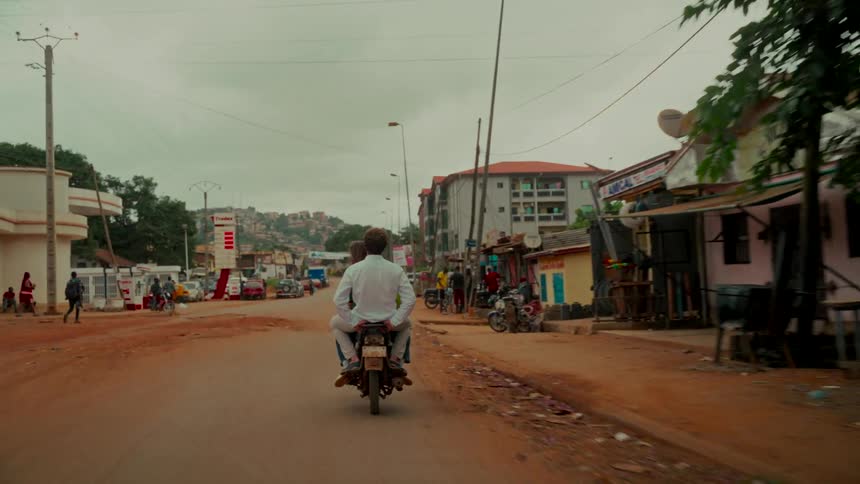A rear tracking shot follows two people riding a motorcycle down a dusty road through a town in a developing country