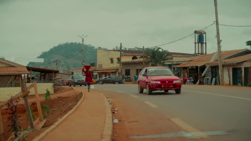 A person wearing red balances a basin on their head while walking along a road in a small African town with hills in the background