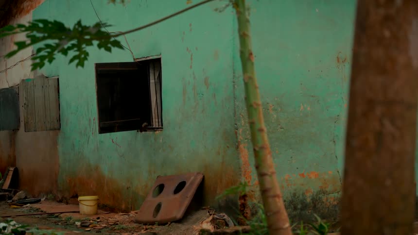 A static exterior shot of a weathered, green-painted house in a rural or impoverished setting