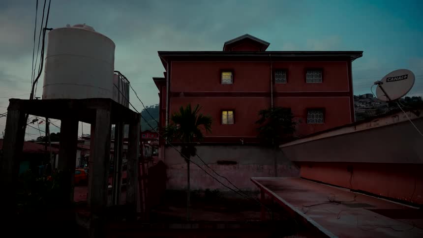 A red residential building with glowing windows sits next to a water tower at dusk in a quiet hillside town