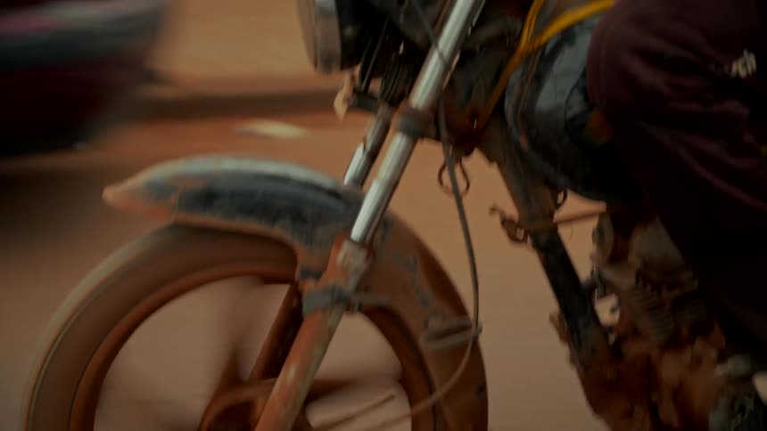 Detailed slow-motion shot pans up from the muddy spinning wheel of a motorcycle to the driver navigating a dusty road