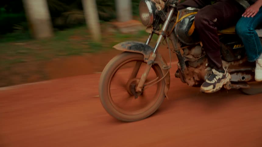 A close-up tracking shot follows the front wheel of a muddy motorcycle as it drives down a red dirt road in the countryside