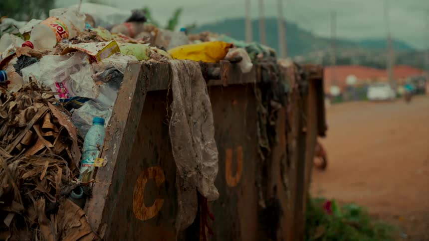 An overflowing dumpster filled with garbage and plastic waste stands on the side of a dirt road as traffic passes by