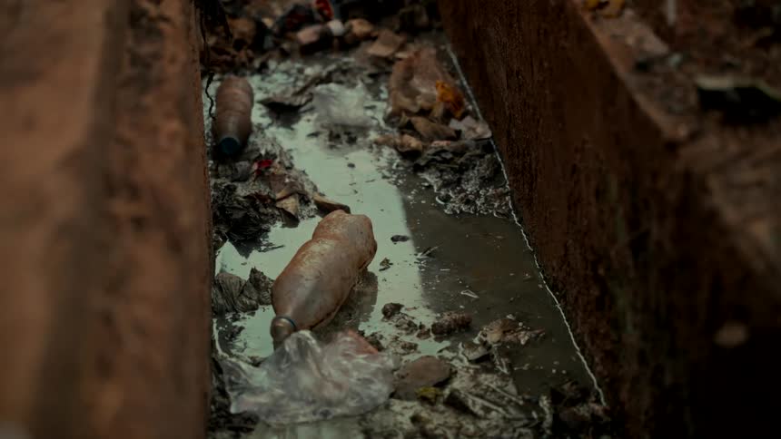 Plastic pollution and garbage clog a narrow drainage channel filled with dirty water