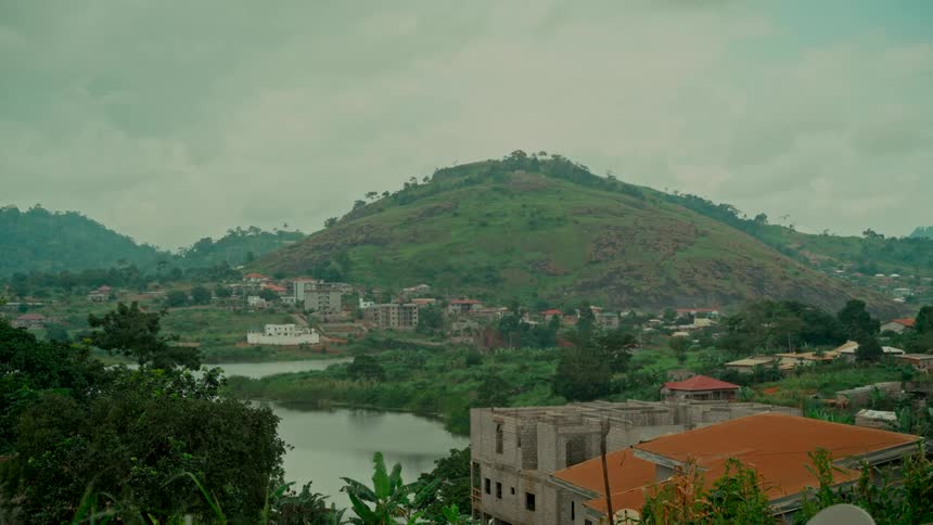 A wide, static shot of a verdant mountain towering over a small town and a serene lake under an overcast sky