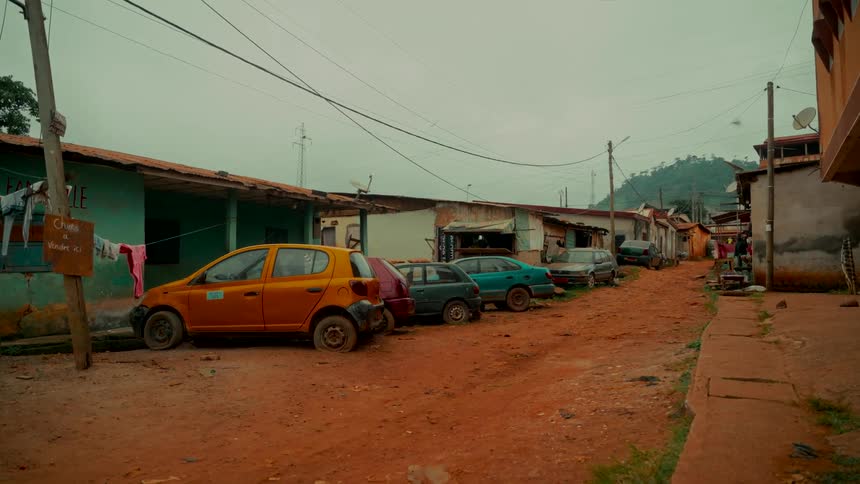Old cars are parked on a red dirt road running through a rural village in a developing country on a cloudy day