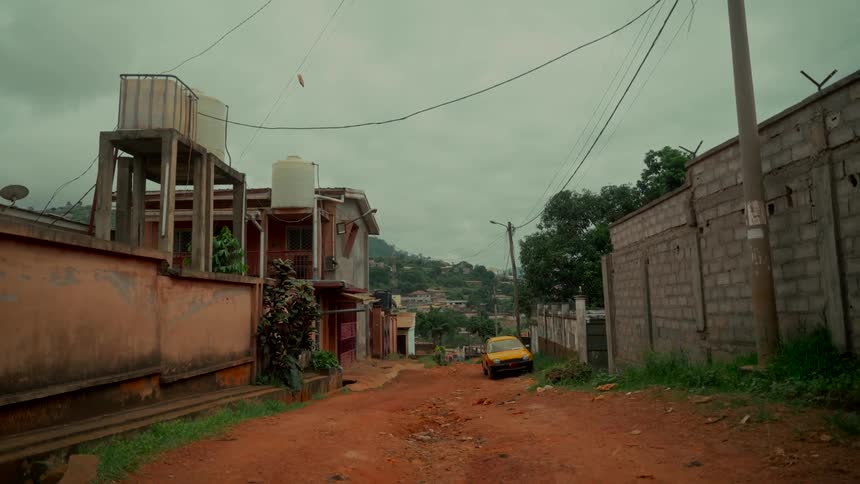 A yellow car sits parked on a quiet, unpaved red dirt street in a hilly town on an overcast day