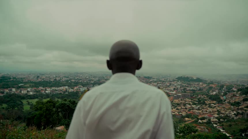A man in a white uniform shirt stands on a hill overlooking a sprawling cityscape on a cloudy, overcast day
