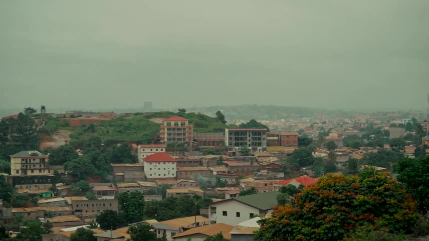 A slow pan reveals a sprawling cityscape of buildings nestled amongst green, rolling hills on a hazy, overcast day