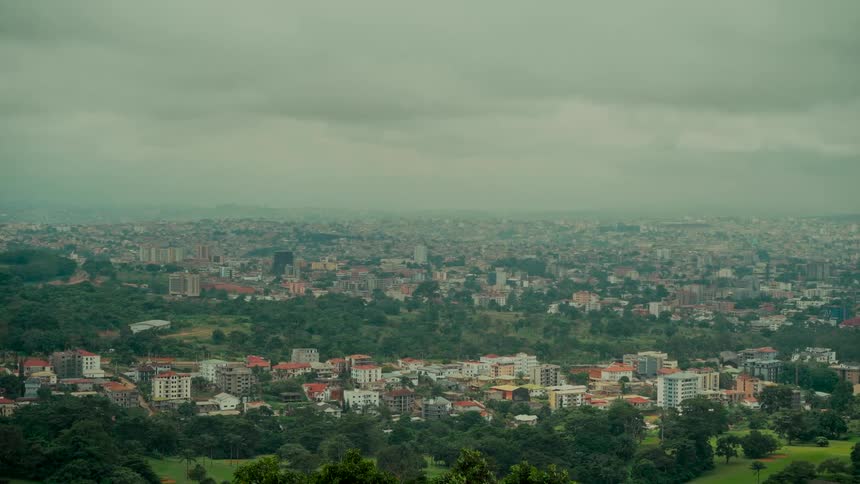 A wide, static view of a sprawling cityscape with lush greenery under a hazy, overcast sky