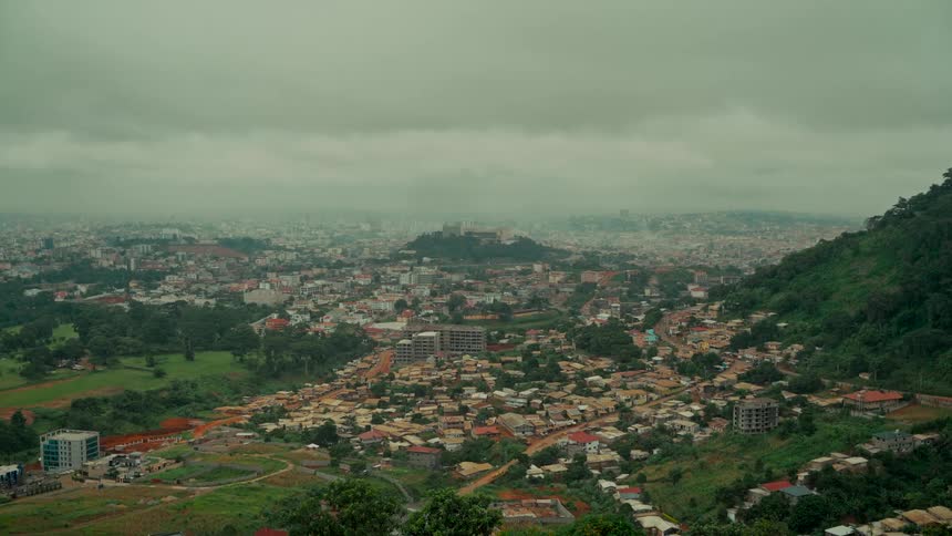 A wide shot of the sprawling city of Yaoundé, Cameroon, nestled among green hills under a cloudy, atmospheric sky