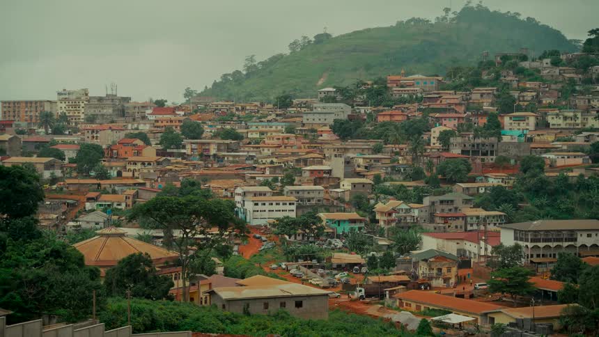 A wide, static view of a colorful city nestled on a lush, green hillside under an overcast sky