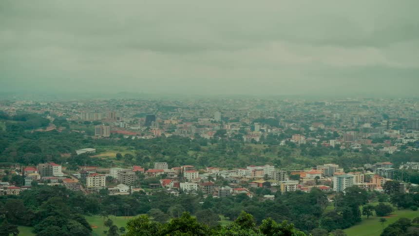 A wide panoramic view of a sprawling cityscape nestled among lush green trees on a cloudy and hazy day