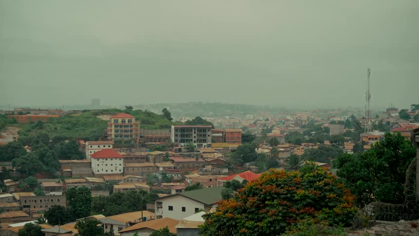A wide, static shot of a sprawling African city with lush green hills on a hazy, overcast day