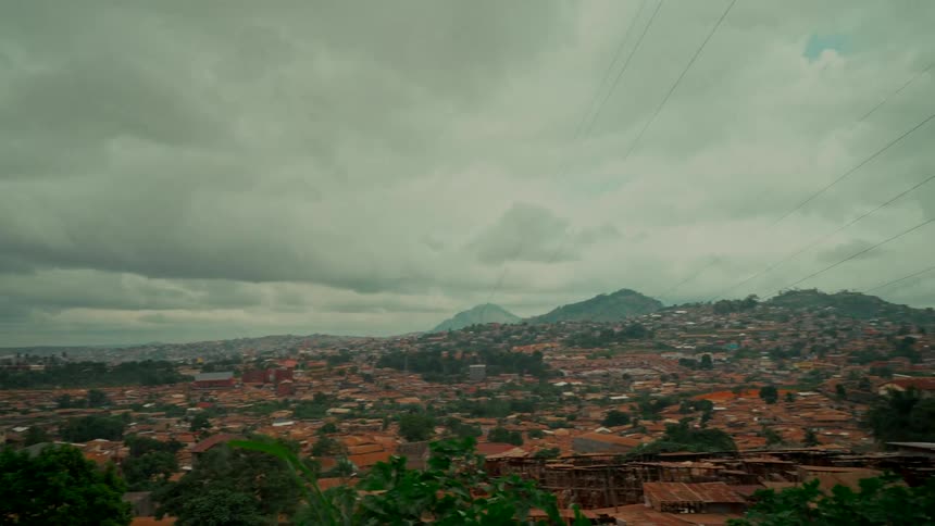 A sweeping panoramic view captures a sprawling city nestled among green hills under an overcast sky