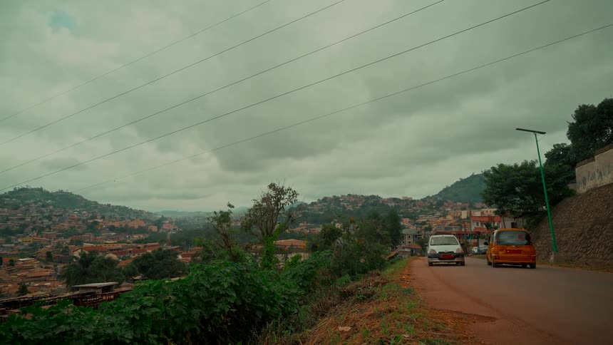 Cars drive along a hillside road overlooking a densely populated city in a valley under a cloudy, grey sky