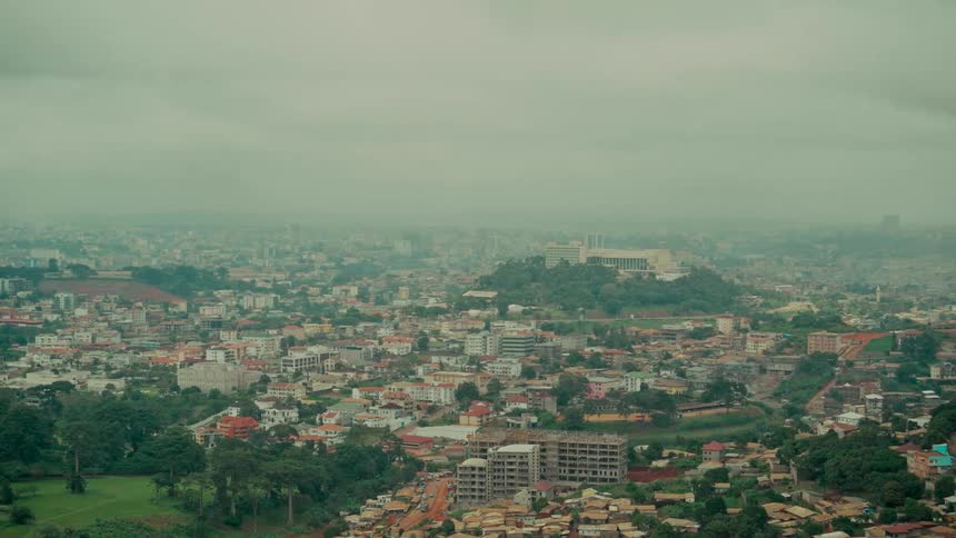 A wide aerial pan over the sprawling city of Yaounde, Cameroon, built across rolling green hills on a hazy, overcast day