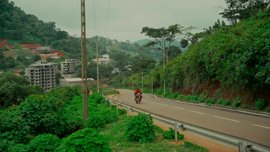 Two people on a motorcycle drive along a winding road through a lush green valley with buildings under construction in the distance