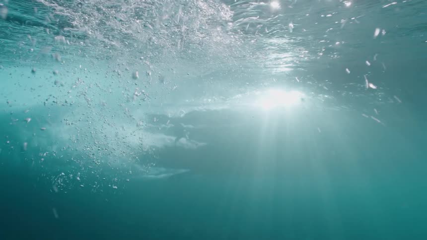 An epic slow-motion underwater view captures a powerful ocean wave as it breaks, sending bubbles through the sunlight-drenched water