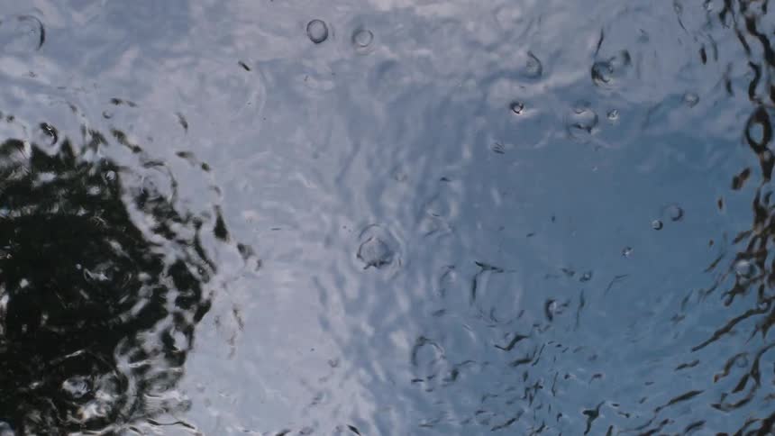 Raindrops fall in slow motion onto the surface of the water, creating abstract patterns and reflecting a cloudy sky