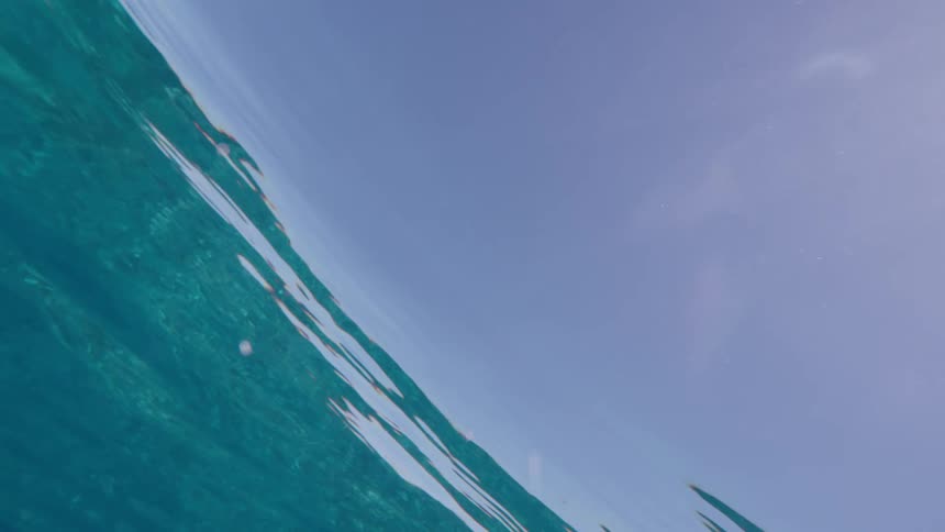 An immersive underwater view looking up at the gently rippling surface of clear blue ocean water on a sunny day