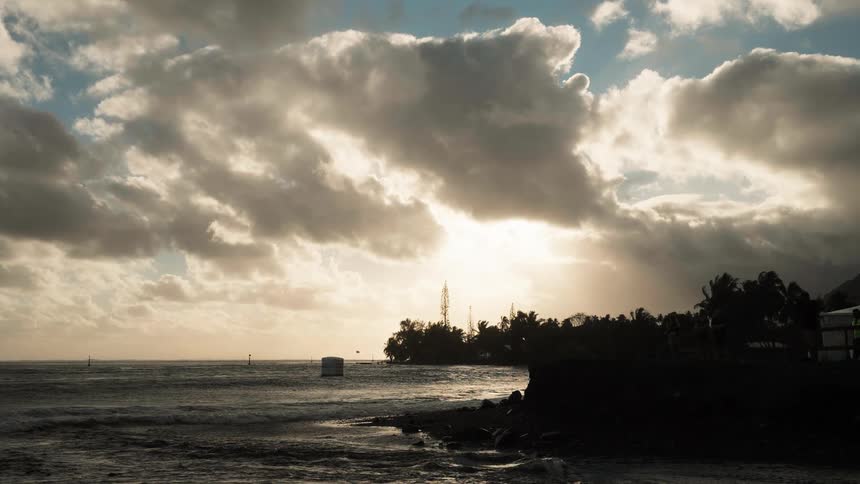 Dramatic clouds part for the setting sun over a silhouetted tropical coastline with gentle waves