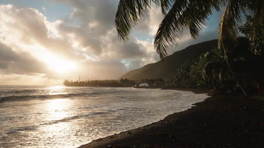 Gentle waves roll onto a black sand beach as the golden sun sets behind a mountainous tropical island