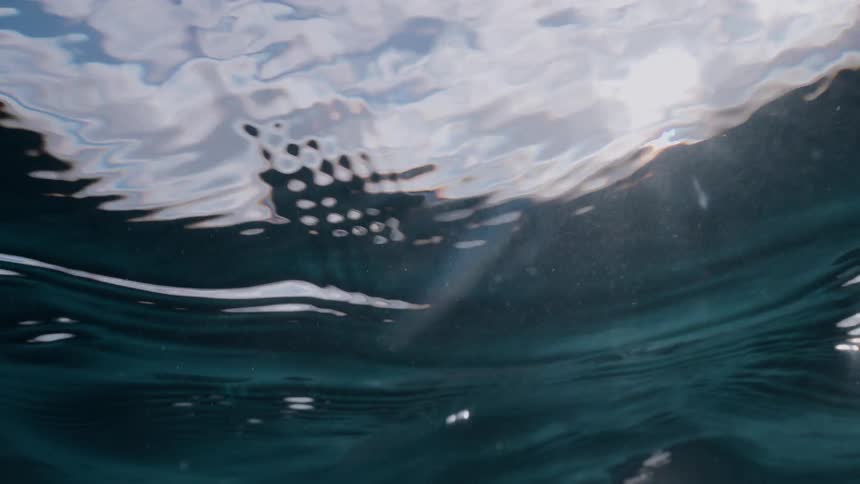 Sunlight filters through the deep blue ocean surface, creating abstract patterns and serene light rays as seen from below