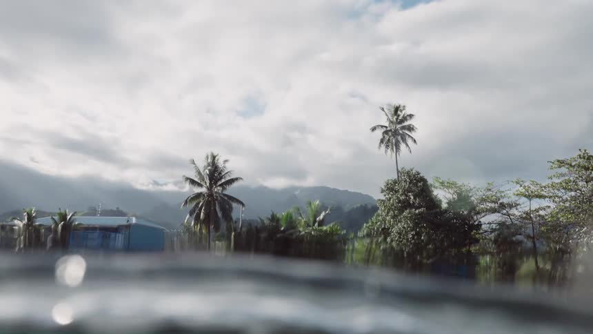 A slow-motion over-under shot reveals a tropical shoreline in Tahiti with colorful homes and palm trees from beneath the ocean surface