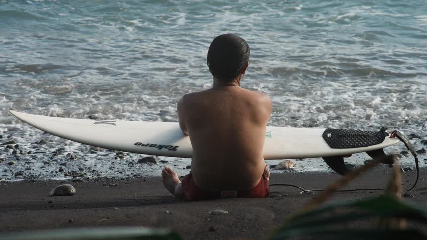 A surfer with his back to the camera sits on a black sand beach with his surfboard, watching the ocean waves roll in