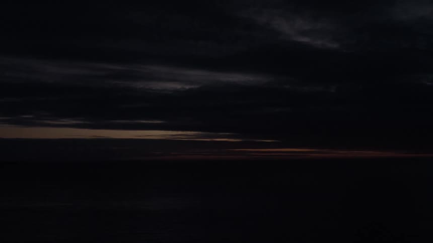 A lone red beacon flashes intermittently on the dark ocean horizon under a cloudy sky at twilight