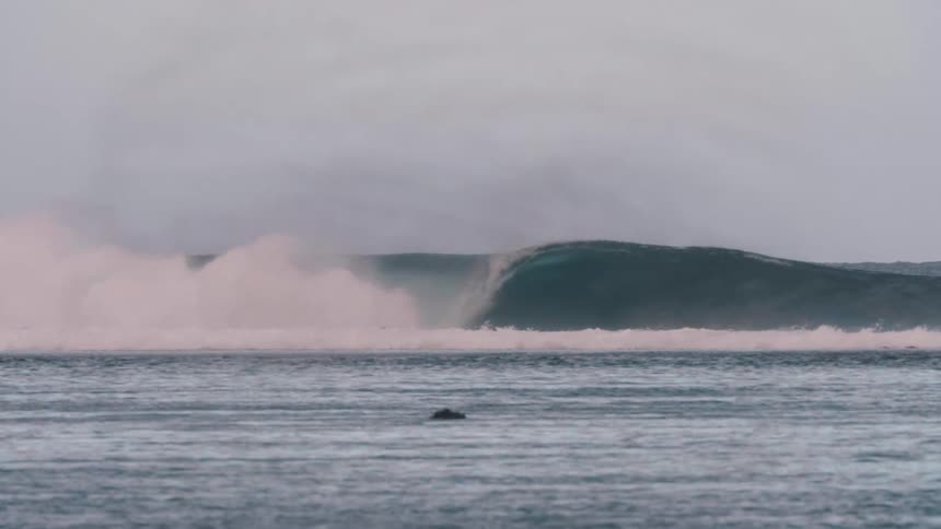 A powerful ocean wave barrels across the water in slow motion on a misty, overcast day