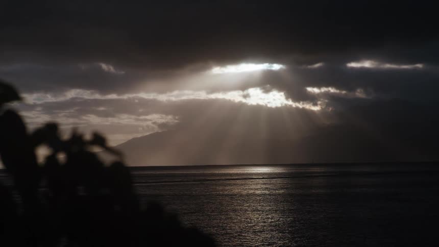 Ethereal sun rays pierce through a moody, dark sky, illuminating the ocean's surface near a distant island