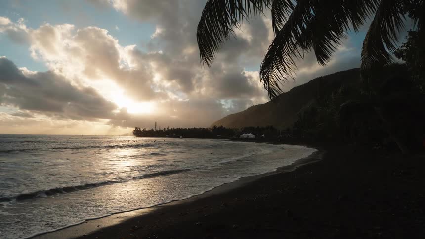 Golden sunset light reflects on the gentle waves of a tropical black sand beach, framed by palm trees and a lush mountain