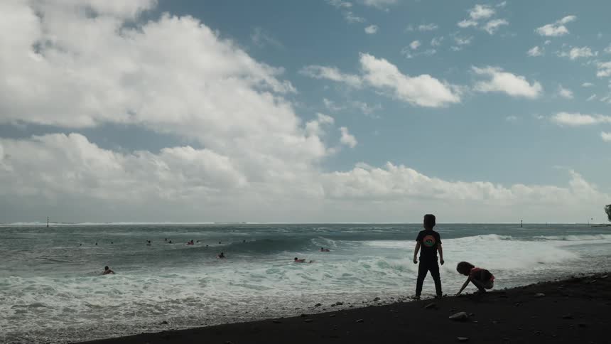 Two children play on the shore of a black sand beach as surfers wait for waves in the ocean