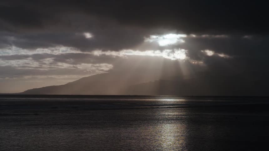 Sun rays break through dramatic, dark clouds, casting a golden light on the ocean and a distant island
