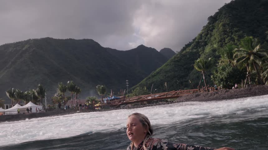 Slow motion reveals a young female surfer paddling on her board with a lush tropical mountain island in the background