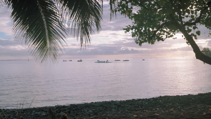 A serene view from a tropical beach of several outrigger canoes paddling on the calm ocean at sunset