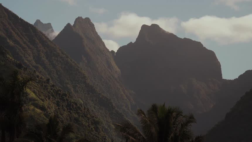 Sunlight illuminates the rugged peaks of a verdant mountain range in a tranquil, tropical setting